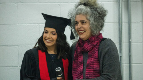 uas mulheres sorriem para a foto. Uma, mulher negra de cabelos trançados longos, veste uma beca e capelo enquanto segura um canudo. A outra mulher branca de cabelos grisalhos veste uma blusa cinza e cachecol rosa ao mesmo tempo em que abraça a outra.