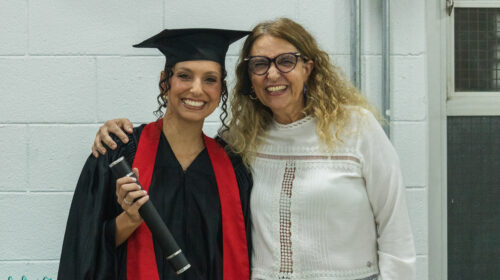 Duas mulheres brancas sorriem para a foto. Uma veste beca e capelo preto a outra uma blusa branca e óculos.