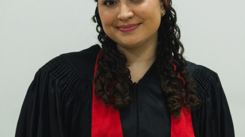 Formanda usandando beca preta, faixa vermelha e capelo preto. Ela é branca, possui cabelo cacheado escuro e está sorrindo discretamente.