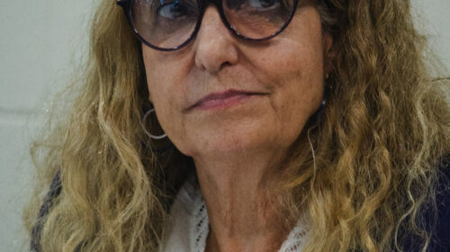 Mulher branca mais velha loira olha para algo extraquadro. Ela veste uma roupa branca e usa óculos.