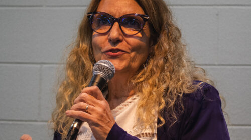 Mulher branca mais velha loira olha para algo extraquadro. Ela veste uma roupa branca e usa óculos. Está falando ao microfone.