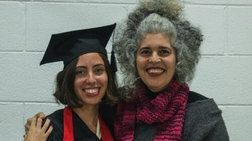 Duas mulheres brancas sorriem para a foto, uma usa trajes tipicos de uma colação de grau a outra usa uma blusa cinza e cachecol.