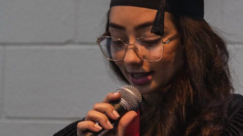 Mulher jovem fala ao microfone. Ela usa beca, capelo e faixa vermelha.