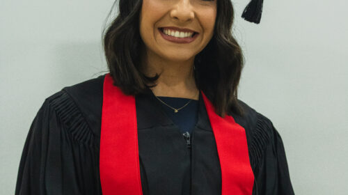 Formanda usando beca, capelo preto e faixa vermelha. Ela é negra de pele clara, possui cabelos com ondas escuro e sorri para a foto com as mãos próximas a barriga.