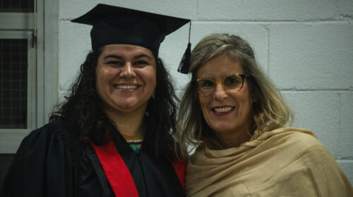Duas mulheres sorriem para a foto. O plano é próximo. Uma veste roupas típicas de uma colação de grau, capelo, beca e faixa vermelha. A outra usa um lenço bege sobre os seus ombros.