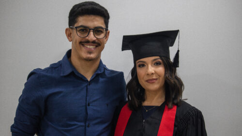 Homem e mulher sorriem discretamente para a foto. A mulher usa beca, capelo e faixa vermelha. O homem usa uma camisa azul de botão. O fundo é branco a iluminação é artificial.