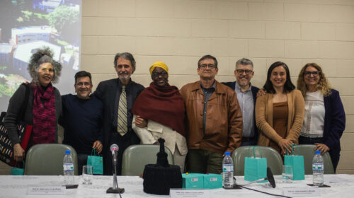 Oito pessoas, quatro mulheres e quatro homens, estão em pé atrás de uma mesa coberta por uma toalha branca, posando para uma foto de grupo. Da esquerda para a direita: uma mulher com cabelo grisalho e cacheado, um homem de óculos e suéter, um homem com cabelos grisalhos e barba, uma mulher negra com turbante amarelo e um lenço bordô sobre os ombros, um homem de jaqueta marrom, um homem de óculos e barba grisalha, uma mulher com blusa e casaco em tons de marrom e, por último, uma mulher de óculos com blusa branca e casaco azul. À frente da mesa, há garrafas de água, sacolas de presente azuis e plaquinhas com nomes, em frente a uma tela com a projeção de uma imagem com edifícios.