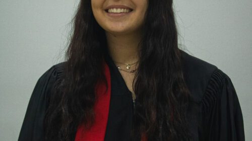 Formanda usando capelo, beca e faixa vermelha. Ela possui cabelo cacheados, é branca e está sorrindo.