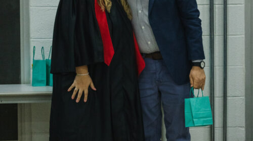 Duas pessoas sorriem para a foto. O homem veste um terno zul e a mulher uma beca e capelo preto.