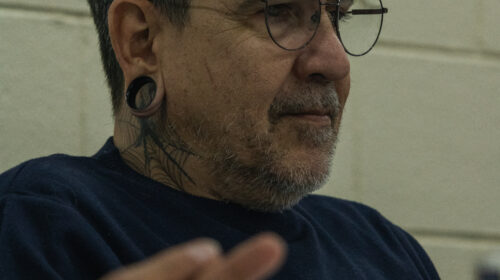 Homem branco de óculos, roupa azul e alargador está de perfil. Em primeiro plano, desfocado, é possível ver canudos.