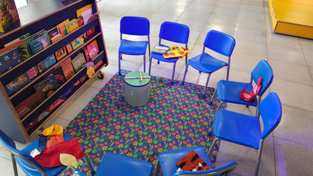 Fotografia que mostra cadeiras pequenas (em tamanho adequado para crianças) na cor azul posicionadas de modo a formar um meio círculo. Há brinquedos nas cadeiras e à frente das cadeiras, há uma estante (também com altura adequadas para crianças) com livros. O ambiente é bem iluminado e bem colorido. Fim da descrição.