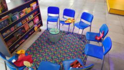 Fotografia que mostra cadeiras pequenas (em tamanho adequado para crianças) na cor azul posicionadas de modo a formar um meio círculo. Há brinquedos nas cadeiras e à frente das cadeiras, há uma estante (também com altura adequadas para crianças) com livros.
