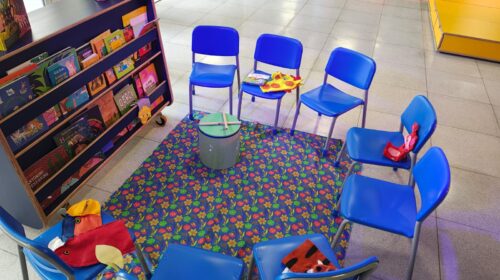 Fotografia que mostra cadeiras pequenas (em tamanho adequado para crianças) na cor azul posicionadas de modo a formar um meio círculo. Há brinquedos nas cadeiras e à frente das cadeiras, há uma estante (também com altura adequadas para crianças) com livros.
