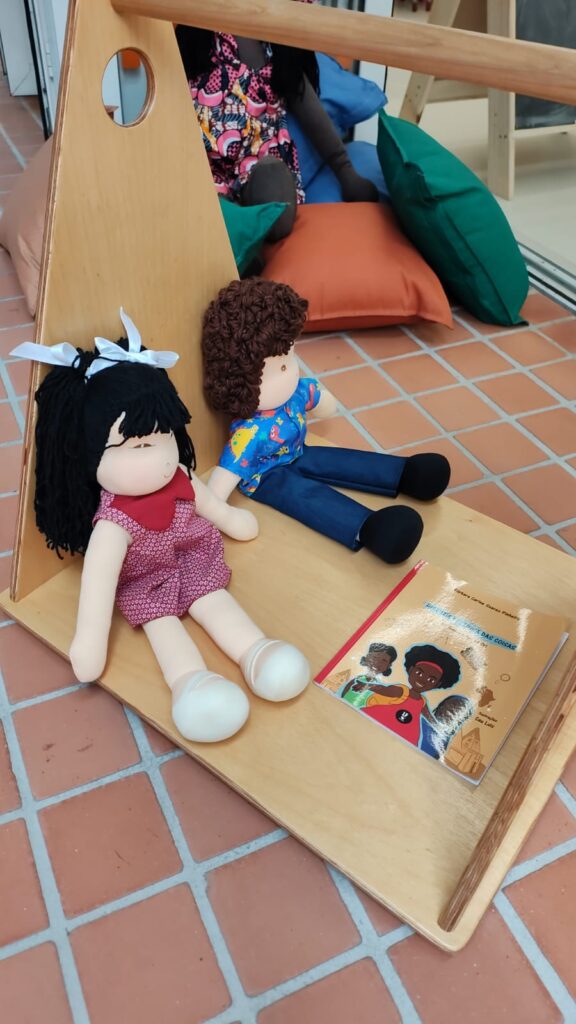 Fotografia de dois bonecos de pano e um livro infantil. O ambiente é cercado por brinquedos e bem iluminado. Fim da descrição.