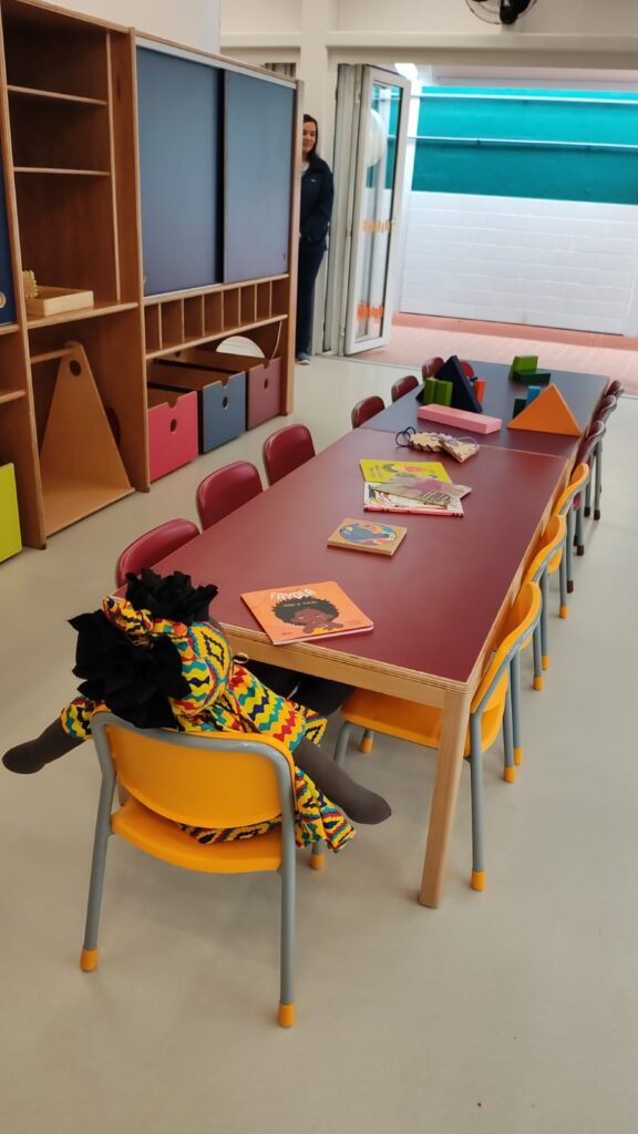 Fotografia de uma sala infantil. O ambiente é parcialmente iluminado, há mesas e cadeiras em tamanho adequado para crianças e bonecos de pano. Fim da descrição.
