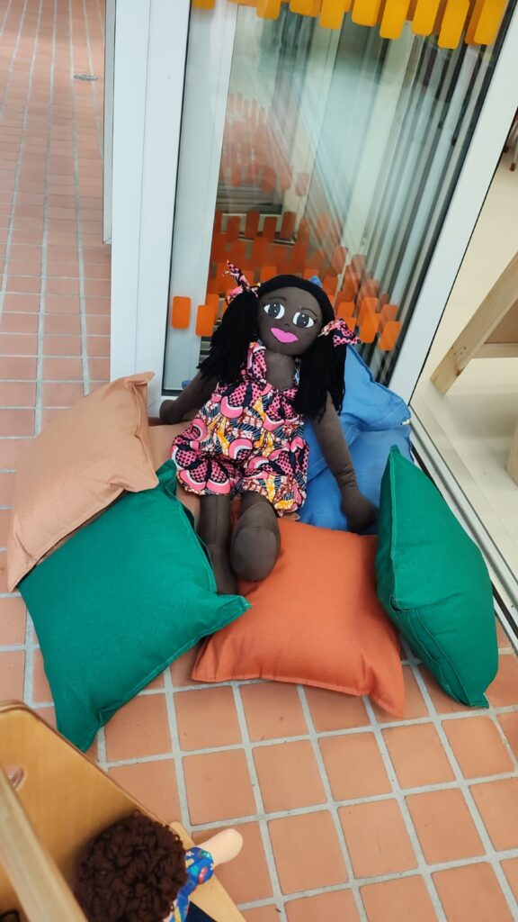 Fotografia de boneca de pano. Ela está posicionada em cima de almofadas em um ambiente que parece ser voltado para o público infantil. Fim da descrição.