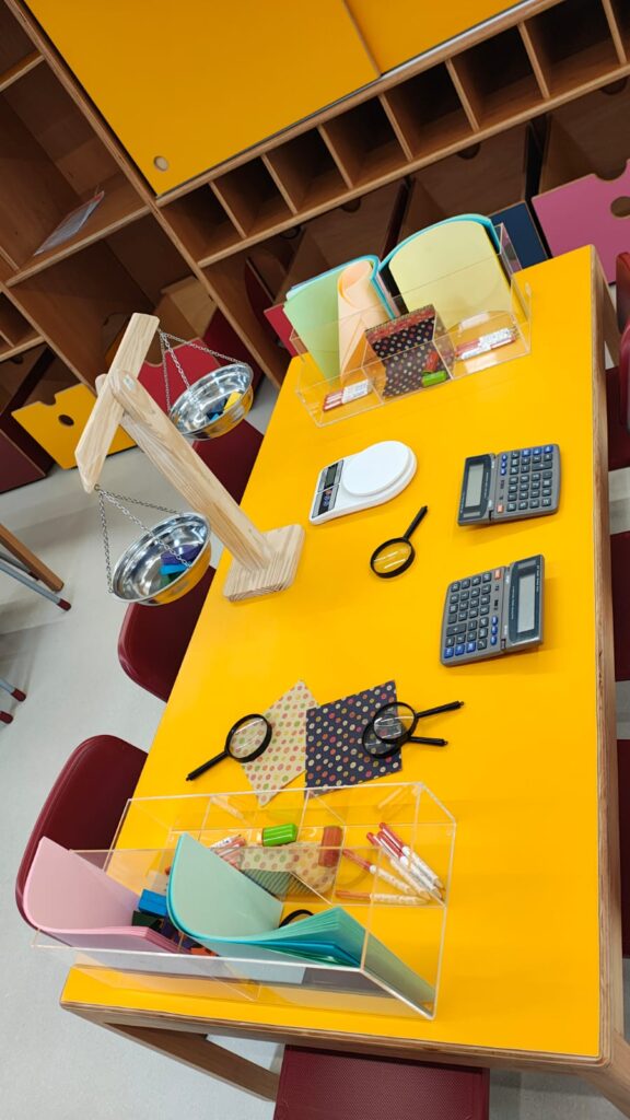 Fotografia de uma mesa amarela com cadeiras vermelhas. Em cima da mesa há calculadoras, lupas e materiais para escrita e desenho. O ambiente é colorido e parece ser um espaço infantil. Fim da descrição.