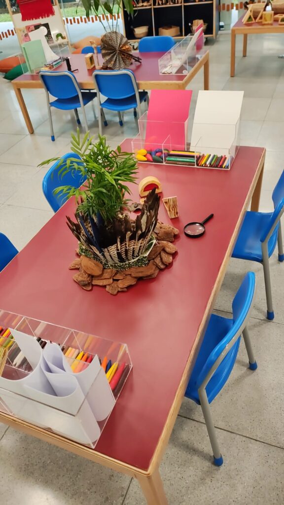 Fotografia que mostra espaço infantil. Há mesas e cadeiras. Em cima das mesas, há materiais para escrita e desenho, lupas e plantas. Fim da descrição. 