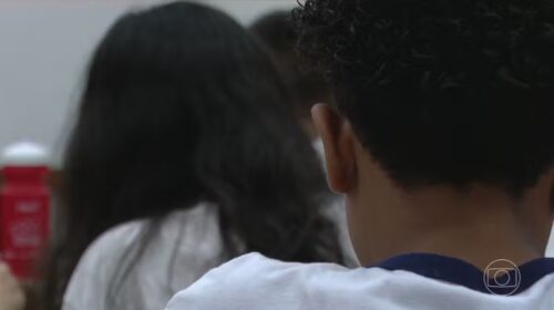 Fotografia que mostra duas crianças. As duas estão de costas para a câmera, que só captura parte do pescoço de da cabeça delas. O ambiente parece ser uma sala de aula. Fim da descrição.