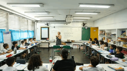 Imagem que mostra sala de aula. As carteiras dos alunos formam um semicírculo e há uma professora ao centro. A sala é repleta de livros e cartazes coloridos. Ao fundo, há uma lousa branca com escritas em preto. Fim da descrição.