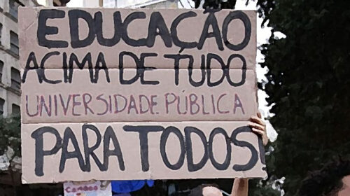 Fotografia que mostra um cartaz de manifestação com a escrita "Educação acima de tudo, universidade pública para todos", escrita em letras grandes. O fundo parece ser uma rua com manifestantes, durante o dia. Fim da descrição.