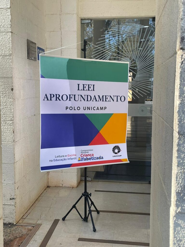 Fotografia que mostra um banner colorido em um ambiente aberto e bem iluminado. No banner, há a escrita "LEEI Aprofundamento Polo Unicamp". Ao fundo, há paredes claras e uma porta de vidro. Fim da descrição.
