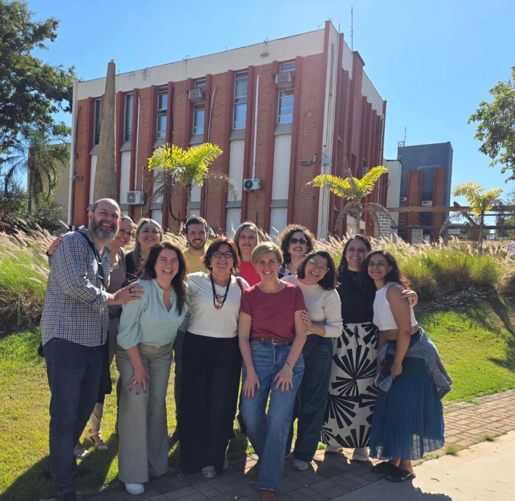 Fotografias que mostra um grupo de pessoas posando e sorrindo na frente de um prédio institucional. O ambiente é aberto, há grama, árvores e está de dia. O grupo é composto por 10 mulheres e 2 homens. Fim da descrição.