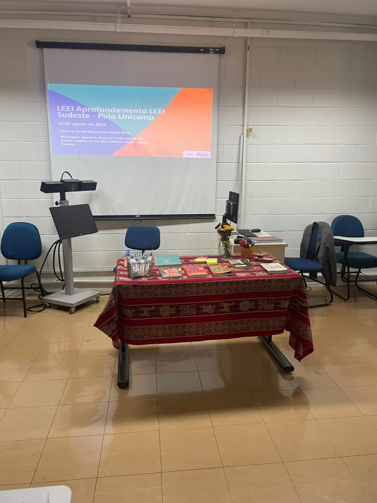Fotografia que mostra uma sala com uma mesa com comidas e um projetor com a escrita "LEEI Aprofundamento LEEI Sudeste - Polo Unicamp". O ambiente é fechado e bem iluminado, com predominância de cores claras. Fim da descrição.
