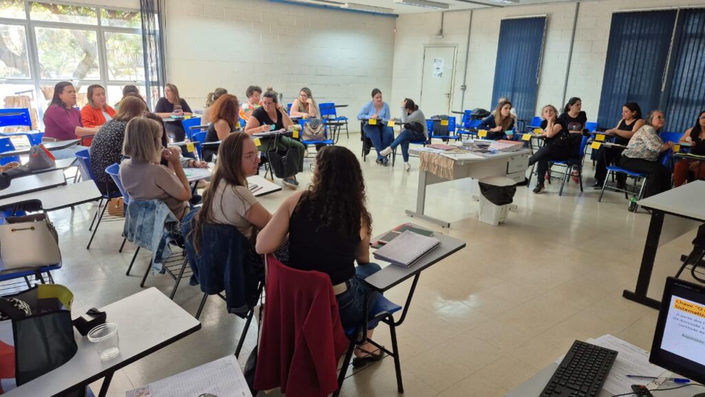Fotografia que mostra várias mulheres adultas em uma sala de aula. O ambiente tem cores claras e janelas grandes, por onde entra iluminação natural. Fim da descrição.