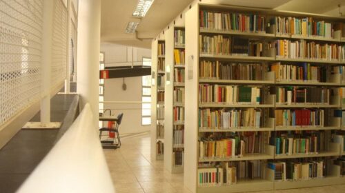 Fotografia que mostra interior da Biblioteca Prof. Joel Martins, da Faculdade de Educação da Unicamp. O ambiente é bem iluminado e há estantes com livros. O piso, as paredes e o teto são claros. Fim da descrição.