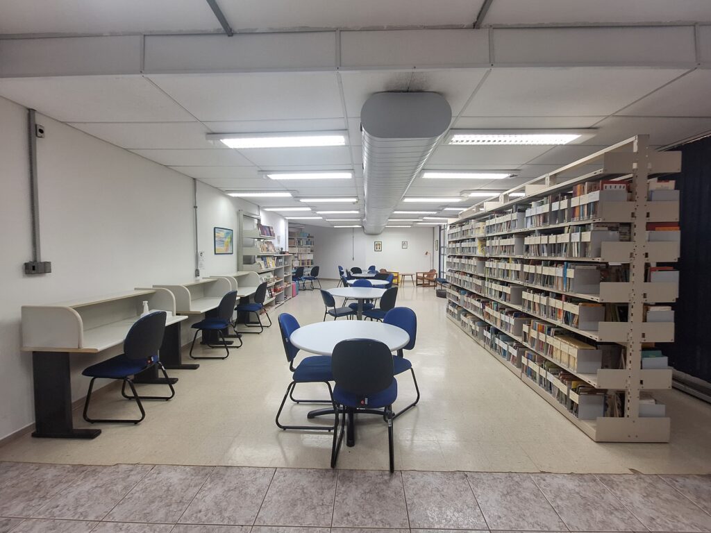 Fotografia que mostra o interior da Biblioteca Joel Martins, da Faculdade de Educação da Unicamp. As paredes, o teto e o piso são claros e há estantes com livros à direita. À esquerda, há escrivaninhas brancas encostadas na parede e cadeiras azuis à frente. Ao centro, há mesas circulares brancas com cadeiras azuis em volta. O ambiente é amplo e bem iluminado. Fim da descrição.