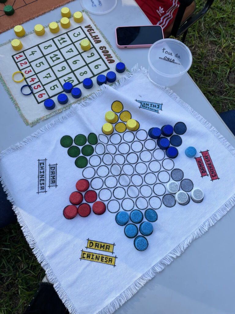 Fotografia tirada durante o dia que mostra jogo dama chinesa em cima de uma mesa. O jogo é fito com materiais reciclados. Fim da descrição.