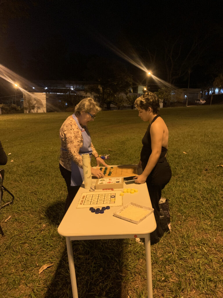 Fotografia que mostra duas mulheres jogando um jogo de tabuleiro. É noite e o ambiente é externo, com grama. Fim da descrição.