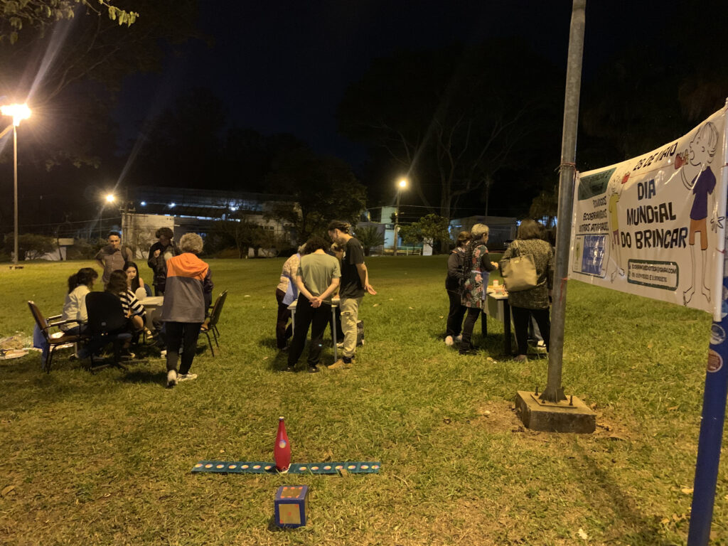 Fotografia que mostra pessoas em ambiente externo interagindo umas com as outras. É noite. Fim da descrição.