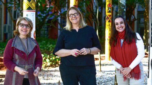 Fotografia que mostra três mulheres posando e sorrindo no Jardim dos Saberes Ancestrais da Faculdade de Educação da Unicamp. Todas vestem roupas elegantes e de inverno. Fim da descrição.