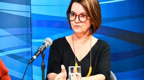 Fotografia que mostra a professora Heloísa Lins sentada á mesa. À frente da professora, há um copo de água e um microfone e, ao fundo, há uma parede azul. Heloísa é uma mulher branca, cabelo castanho curto, usa óculos de grau, veste uma blusa preta e um colar amarelo. Heloísa está falando e gesticulando com as mãos Fim da descrição.