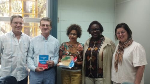 Fotografia que mostra cinco pessoas, dois homens e três mulheres. Todos vestem roupas de trabalho e estão em um ambiente com paredes claras e boas iluminação. Um homem e uma mulher seguram folhetos e livros. Fim da descrição.