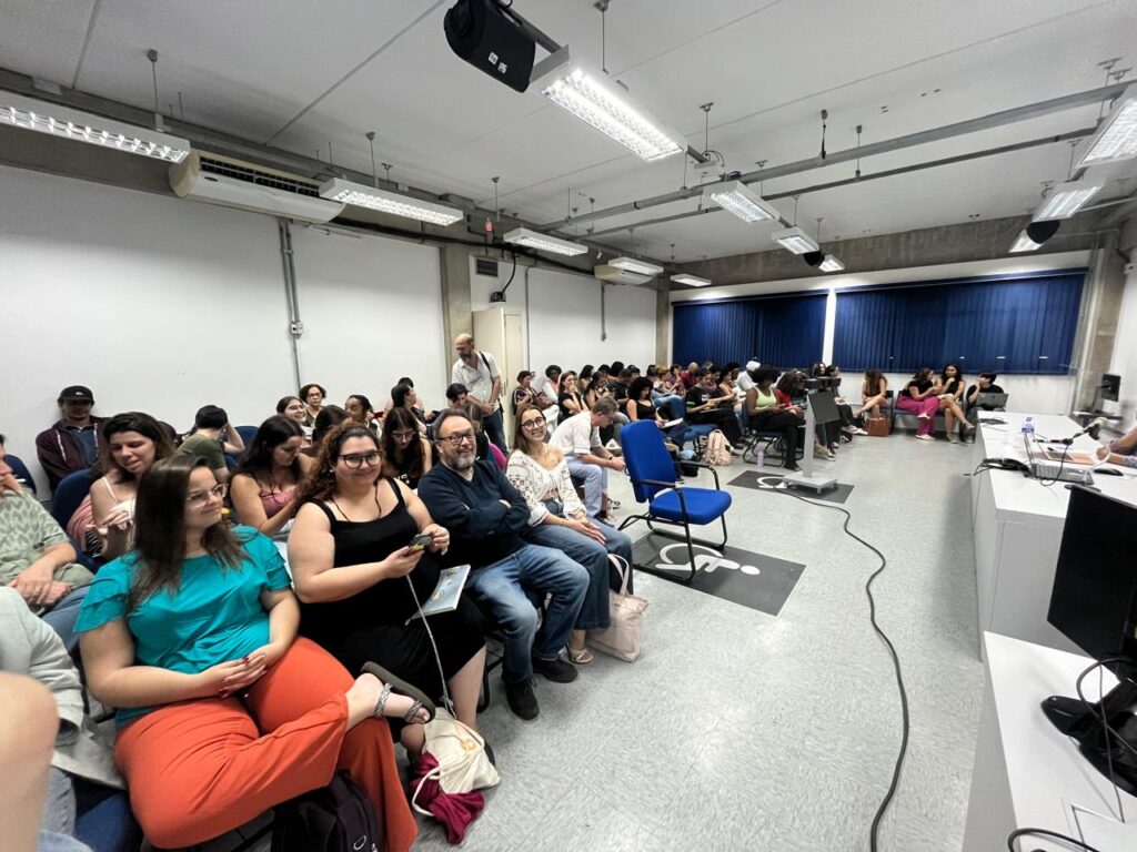 Fotografia que mostra público em ambiente fechado. O ambiente tem paredes, piso e teto claros, janelas cortinas azuis e é bem iluminado e há cadeiras com pessoas sentadas olhando para frente. Fim da descrição.