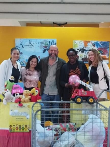 Fotografia que mostra cinco pessoas posando e sorrindo para foto em ambiente fechado e com caixas à sua frente. O grupo é composto por quatro mulheres e um homem e todos vestem roupas formais e de inverno. O fundo é um ambiente fechado; as paredes, o teto e o piso são claros, há janela, porta, bancos, rampa e um fundo amarelo de tecido estendido. À frente do grupo, há duas caixas grandes e decoradas com brinquedos dentro. Fim da descrição.