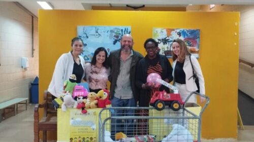 Fotografia que mostra cinco pessoas posando e sorrindo para foto em ambiente fechado e com caixas à sua frente. O grupo é composto por quatro mulheres e um homem e todos vestem roupas formais e de inverno. O fundo é um ambiente fechado; as paredes, o teto e o piso são claros, há janela, porta, bancos, rampa e um fundo amarelo de tecido estendido. À frente do grupo, há duas caixas grandes e decoradas com brinquedos dentro. Fim da descrição.