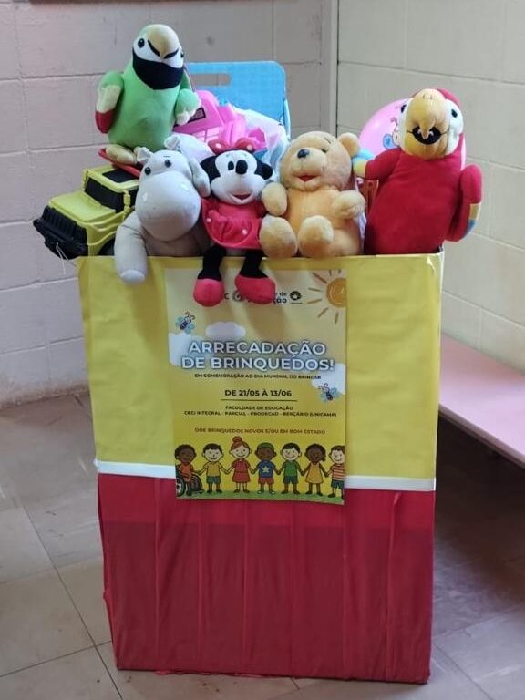 Fotografia que mostra caixa grande e decorada com brinquedos dentro. O ambiente é fechado e tem paredes e piso claros e boa iluminação. A caixa é amarela e vermelha, com laço de cetim branco e um cartaz de campanha colado. O cartaz é amarelo e nele há a escrita "Arrecadação de brinquedos!". Os brinquedos dentro da caixa são animais de pelúcia, bola, carro de brinquedo, entro outros. Fim da descrição.