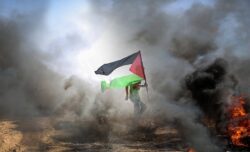 Fotografia colorida. Um homem levanta a bandeira palestina em meio a fumaça. Fim da descrição.