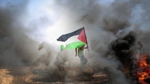 Fotografia colorida. Um homem levanta a bandeira palestina em meio a fumaça. Fim da descrição.