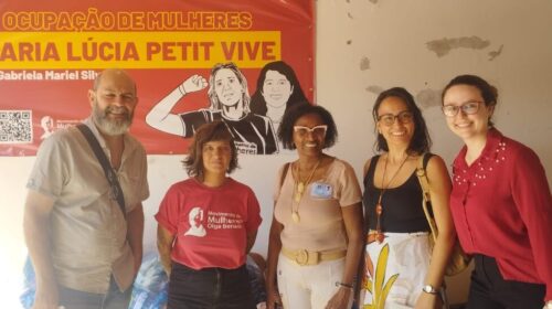 Fotografia que mostra cinco pessoas, uma ao lado da outra, olhando para a câmera. Há quatro mulheres e um homem. O fundo é uma parede branca com um tecido vermelho com a escrita "Ocupação de mulheres Maria Lúcia Petit Vive" em amarelo. Fim da descrição.