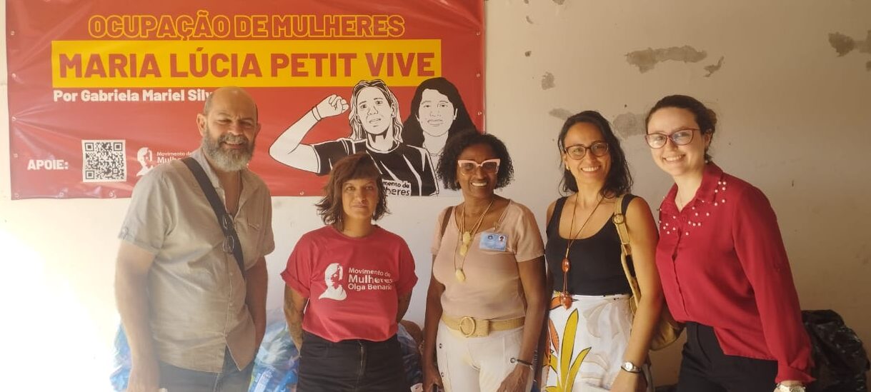 Fotografia que mostra cinco pessoas, uma ao lado da outra, olhando para a câmera. Há quatro mulheres e um homem. O fundo é uma parede branca com um tecido vermelho com a escrita "Ocupação de mulheres Maria Lúcia Petit Vive" em amarelo. Fim da descrição.