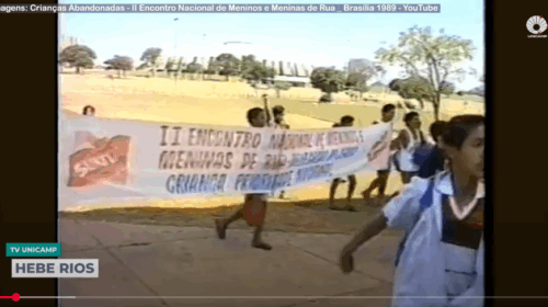 Captura de tela de computador que mostra parte de vídeo no YouTube. Ao centro, há uma imagem do II Encontro Nacional de Meninos e Meninas de Rua, ocorrido em 1989. Abaixo, alinhadas à lateral direita, há duas caixas de texto com as escritas "TV Unicamp" e "Hebe Rios". Fim da descrição.