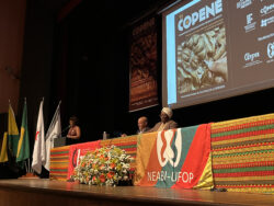 Fotografia que mostra palco em auditório. A fotografia foi tiradas pela visão das primeiras fileiras do lado direito da plateia. Ao centro do palco, há uma mesa comprida coberta por tecidos estampados com padrões geométricos que remetem à cultura africana com as cores vermelho, amarelo, verde e marrom como predominantes; pendurada na mesa há também uma bandeira com a escrita "NEABI-UFOP". Sentados à mesa, há um homem e uma mulher. À esquerda, há uma mulher falando no microfone apoiada em uma tribuna. Todas as pessoas vestem roupas formais. À esquerda, há quatro bandeiras penduradas. Atrás, na parede, está projetada uma página com o nome "COPENE". Fim da descrição.