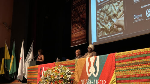 Fotografia que mostra palco em auditório. A fotografia foi tiradas pela visão das primeiras fileiras do lado direito da plateia. Ao centro do palco, há uma mesa comprida coberta por tecidos estampados com padrões geométricos que remetem à cultura africana com as cores vermelho, amarelo, verde e marrom como predominantes; pendurada na mesa há também uma bandeira com a escrita "NEABI-UFOP". Sentados à mesa, há um homem e uma mulher. À esquerda, há uma mulher falando no microfone apoiada em uma tribuna. Todas as pessoas vestem roupas formais. À esquerda, há quatro bandeiras penduradas. Atrás, na parede, está projetada uma página com o nome "COPENE". Fim da descrição.