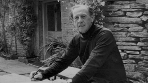 Fotografia em preto e branco que mostra Raymond Williams, homem branco, que veste blusa de frio escura e posa com uma caneta na mão na frente de uma construção que se assemelha a uma casa simples. Fim da descrição.