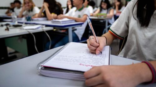 Fotografia que mostra sala de aula. Em primeiro plano, há uma adolescente sentada e escrevendo em seu caderno. Ao fundo, desfocados, há mais adolescentes sentados. Fim da descrição.